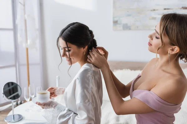Young woman making hairstyle to smiling bride holding coffee in bedroom — Stock Photo
