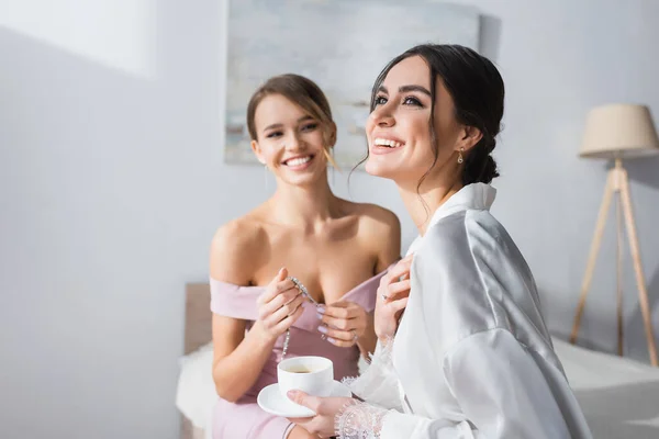 Sposa felice tenendo tazza di caffè e guardando lontano vicino amico con collana su sfondo sfocato — Foto stock