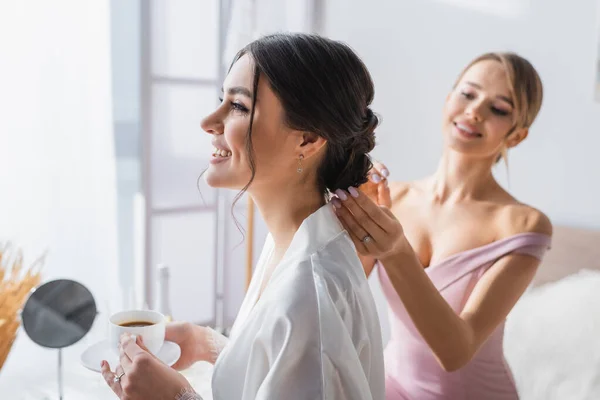 Fröhliche Braut hält Kaffeetasse, während Brautjungfer macht Frisur auf verschwommenem Hintergrund — Stockfoto