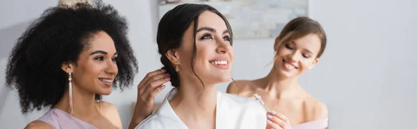 Happy bride looking away while interracial bridesmaids doing hairstyle, banner — Stock Photo
