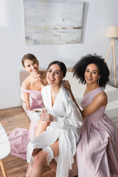 Excited bride in satin robe sitting in bedroom with elegant interracial bridesmaids — Stock Photo
