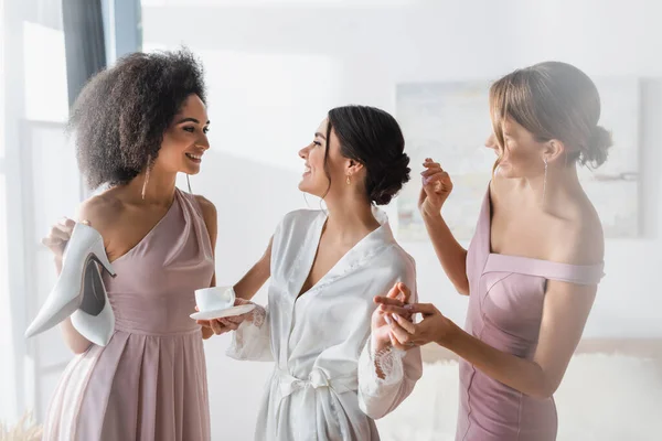 Mujer alegre en bata de seda sonriendo cerca de amigos interracial preparándola para la boda - foto de stock