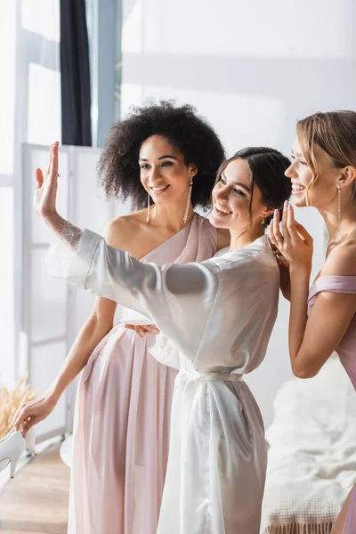 Joyful bride looking at wedding ring on hand near excited multicultural bridesmaids — Stock Photo