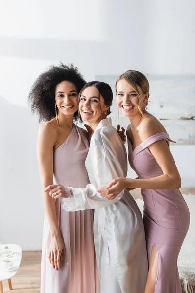 Cheerful bride in satin robe smiling at camera with interracial bridesmaids in bedroom — Stock Photo