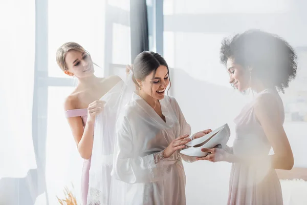 Amazed bride looking at white shoes in hands of african american bridesmaid, blurred foreground — Stock Photo