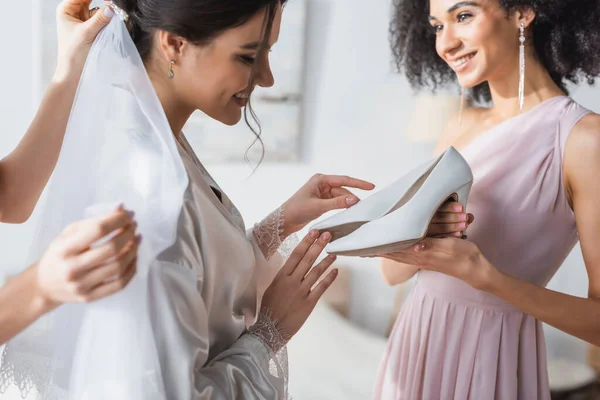 Pleased bride looking at elegant wedding shoes in hands of african american friend — Stock Photo