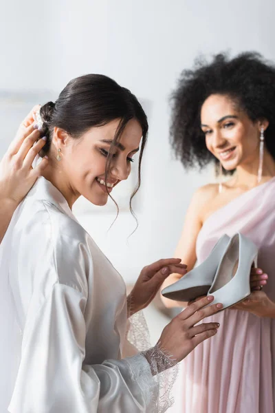 Demoiselles d'honneur interracial faisant coiffure et tenant des chaussures près de la mariée, fond flou — Photo de stock