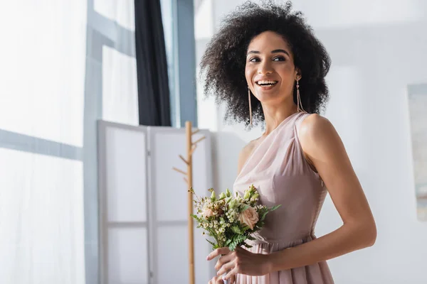 Alegre afro-americana dama de honra olhando embora enquanto segurando buquê de casamento — Fotografia de Stock