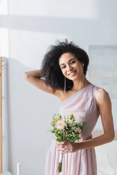 Charmante demoiselle d'honneur afro-américaine touchant les cheveux tout en tenant bouquet de mariage — Photo de stock