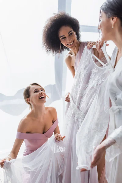 Young woman holding white wedding dress near excited multicultural friends — Stock Photo