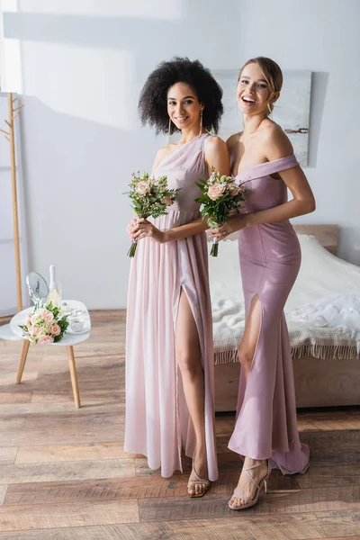 Damas de honra inter-raciais elegantes com buquês de casamento sorrindo para a câmera no quarto — Fotografia de Stock