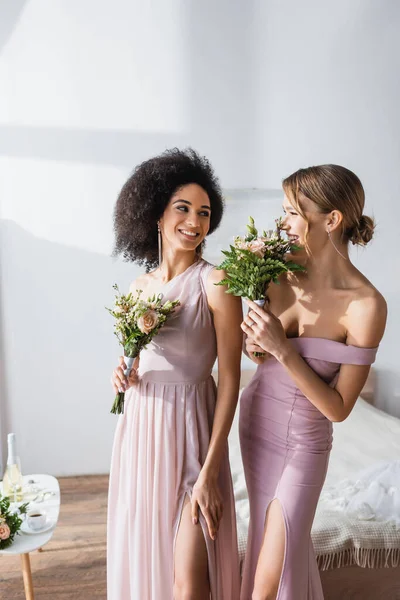 Возбужденные межрасовые подружки невесты улыбаются друг другу, держа свадебные букеты — стоковое фото