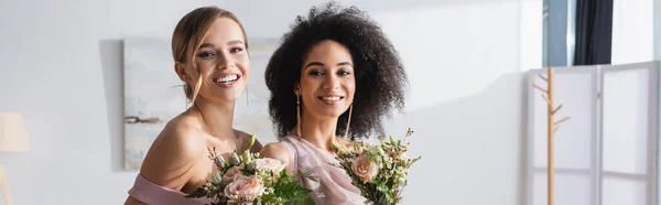 Glücklich gemischtrassige Brautjungfern, die in die Kamera schauen, während sie Hochzeitssträuße, Banner — Stockfoto