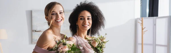 Damas de honor interracial feliz mirando a la cámara mientras sostiene ramos de boda, pancarta - foto de stock