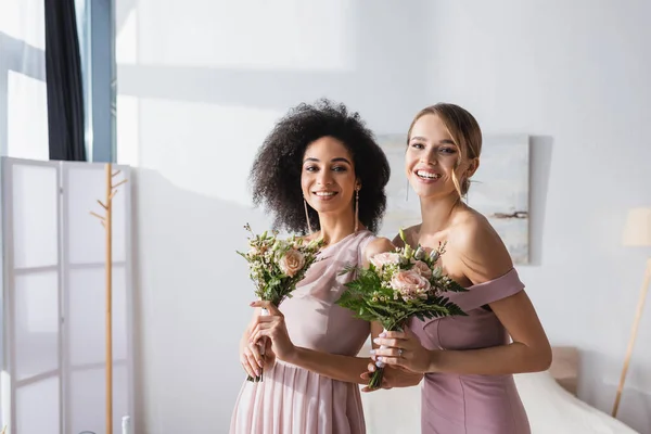 Alegres damas de honor multiculturales sonriendo a la cámara mientras sostiene ramos de boda - foto de stock