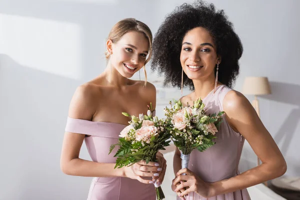 Belas damas de honra multiétnicas segurando buquês de casamento e sorrindo para a câmera — Stock Photo