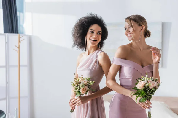 Joyeuses demoiselles d'honneur multiculturelles tenant des bouquets de mariage dans la chambre — Photo de stock