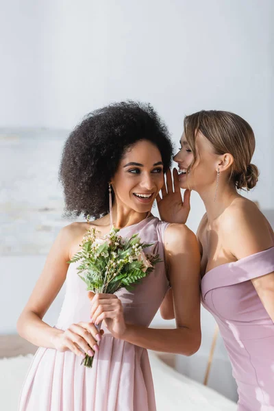 Fröhliche Frau verrät Geheimnis zu lächelnden afrikanisch-amerikanischen Freund mit Brautstrauß — Stockfoto