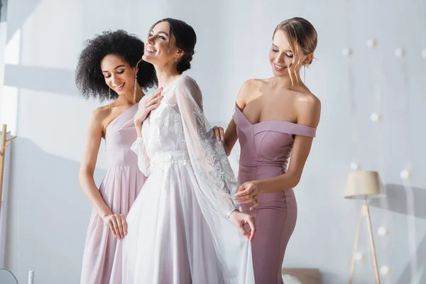 Joyful bride holding hand on chest while multicultural bridesmaids adjusting her dress — Stock Photo