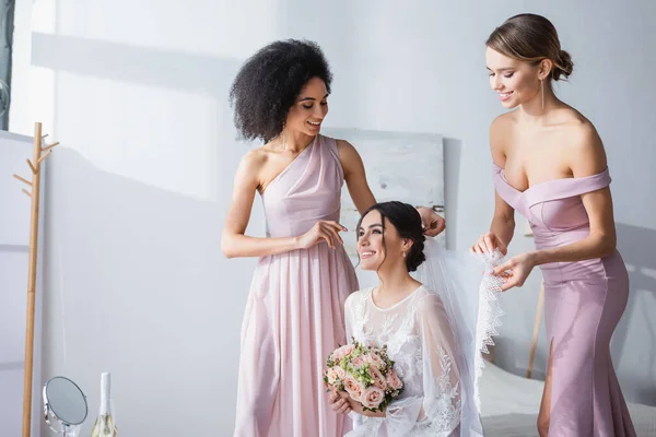 Interracial bridesmaids fixing veil of happy bride sitting in bedroom with wedding bouquet — Stock Photo