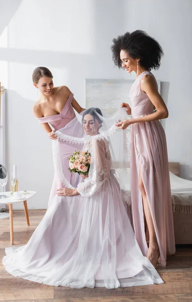 Elegant interracial bridesmaids covering young bride with veil in bedroom — Stock Photo