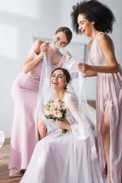 Excitado interracial las mujeres cubriendo overjoyed novia con velo en dormitorio - foto de stock