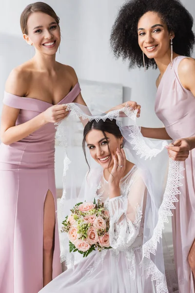 Happy interracial bridesmaids smiling at camera while holding veil over young bride — Stock Photo