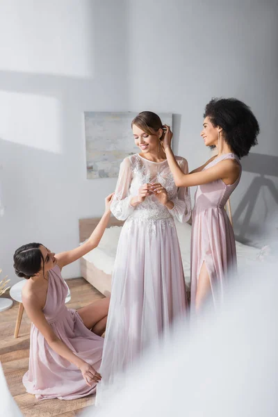 Многокультурные подружки невесты готовят молодую женщину к свадьбе в спальне, размытый передний план — стоковое фото
