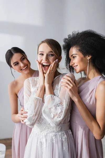 Verblüffte Braut berührt Gesicht beim Blick in die Kamera in der Nähe multikultureller Brautjungfern — Stockfoto