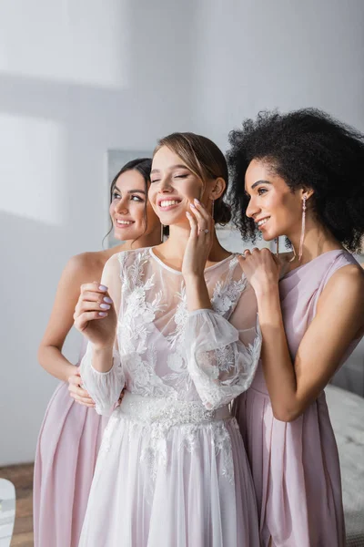 Glückliche Braut mit geschlossenen Augen berührt Gesicht in der Nähe lächelnde interrassische Brautjungfern — Stockfoto