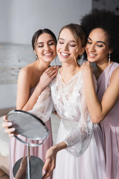 Glückliche interrassische Frauen umarmen lächelnde Braut, die in den Spiegel schaut — Stockfoto