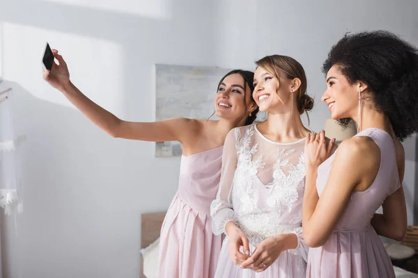 Alegre mujer tomando selfie con novia y africano americano amigo en dormitorio - foto de stock