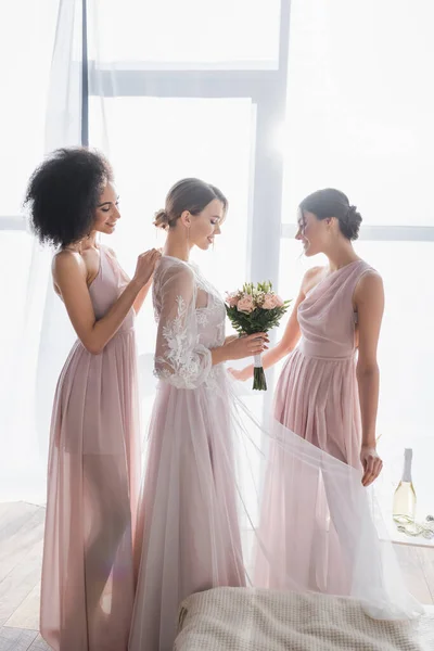 Jolie mariée tenant bouquet de mariage tandis que les demoiselles d'honneur interracial ajuster sa robe le matin ensoleillé — Photo de stock