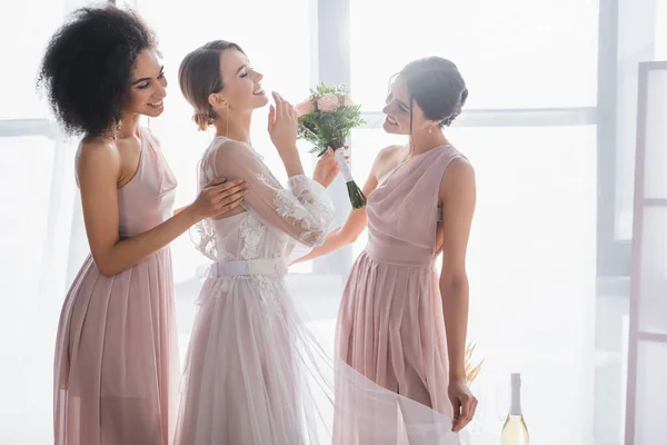 Happy bride holding wedding bouquet and laughing with closed eyes near interracial bridesmaids — Stock Photo