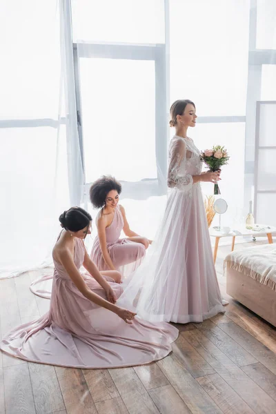 Damas de honor multiculturales ajustando el vestido de novia encantadora en el dormitorio - foto de stock