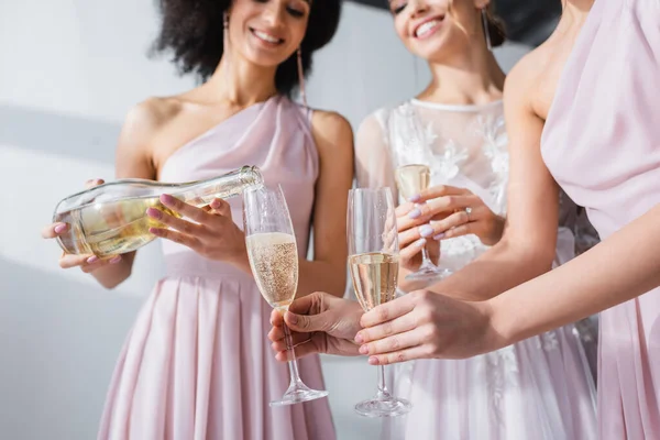 Vista recortada de la mujer afroamericana vertiendo champán en copas de novia y dama de honor, fondo borroso - foto de stock