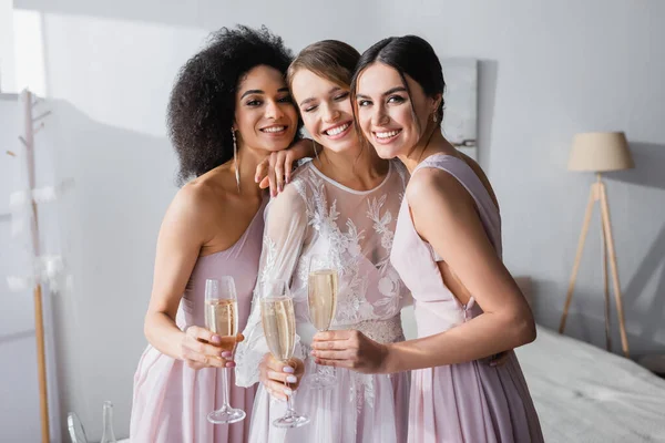 Cheerful interracial bridesmaids holding champagne glasses near happy bride in bedroom — Stock Photo