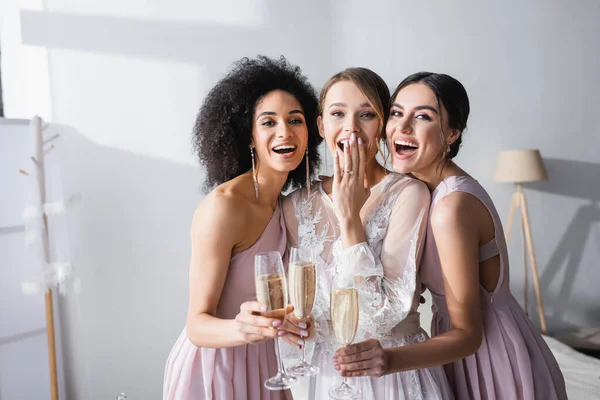 Rindo noiva cobrindo boca com mão perto de damas de honra inter-raciais com copos de champanhe — Fotografia de Stock