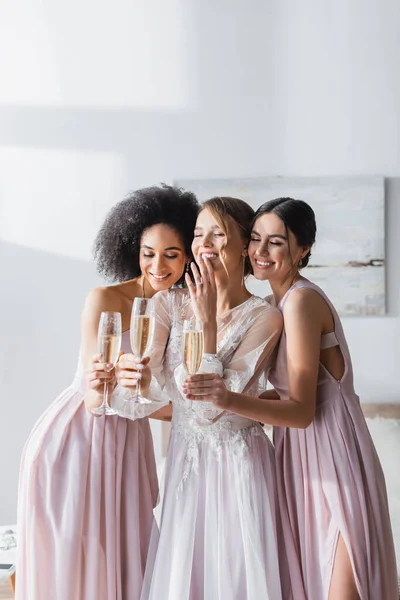 Noiva animado rindo com os olhos fechados perto de damas de honra inter-raciais segurando champanhe — Stock Photo