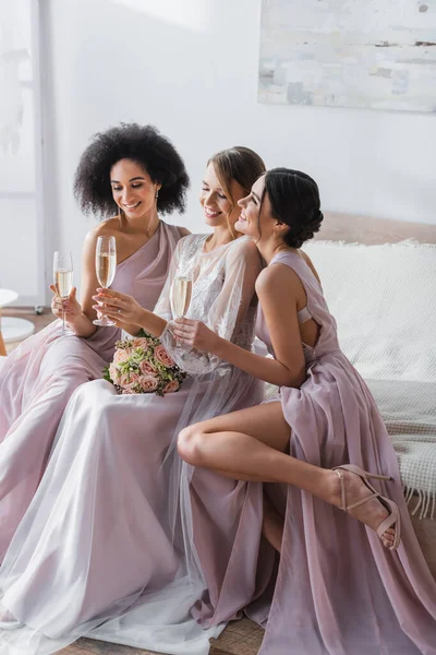 Elegant bride and multicultural bridesmaids sitting with champagne glasses in bedroom — Stock Photo