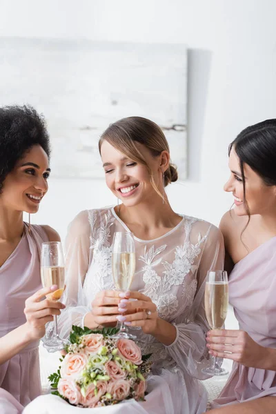Damas de honor multiculturales mirando a la novia feliz mientras sostiene copas de champán - foto de stock