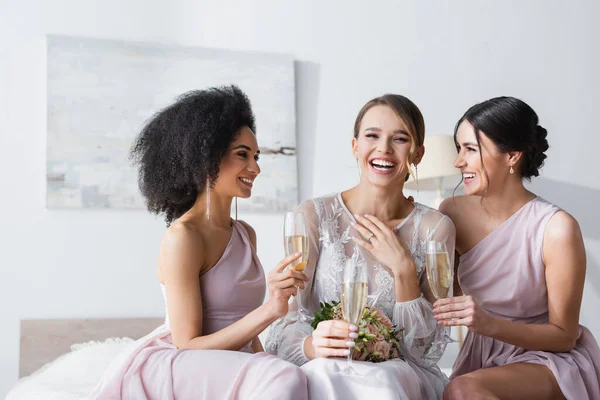 Young bride laughing near interracial friends holding champagne glasses in bedroom — Stock Photo
