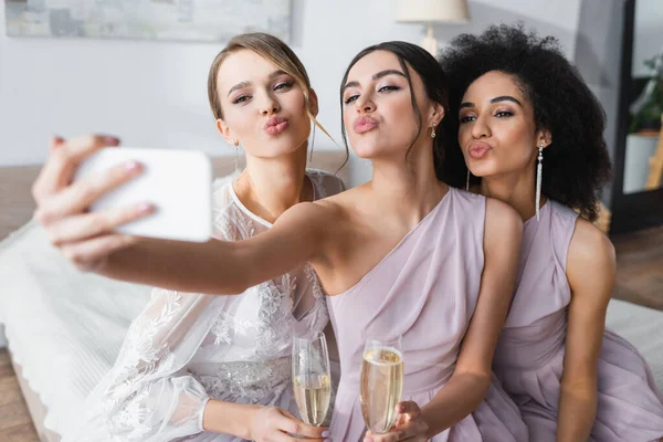 Pretty bride with multicultural bridesmaids blowing air kisses while taking selfie on blurred foreground — Stock Photo