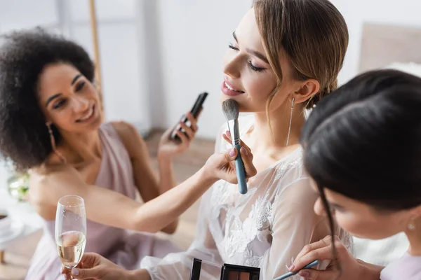 Jovem noiva segurando taça de champanhe enquanto damas de honra multiculturais aplicando maquiagem, fundo borrado — Fotografia de Stock