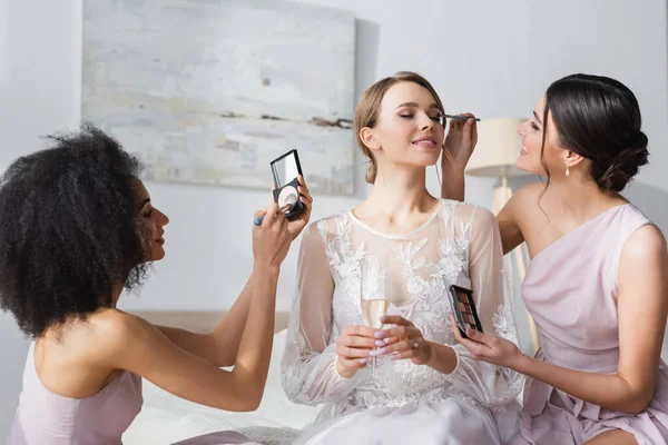 Sposa sorridente che tiene champagne mentre damigelle d'onore interrazziale applicare il trucco — Foto stock