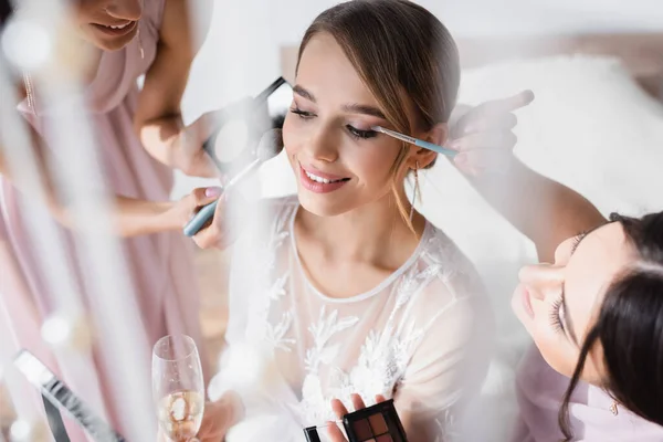 Міжрасові жінки роблять макіяж молодої нареченої, тримаючи келих шампанського на розмитому фоні — стокове фото