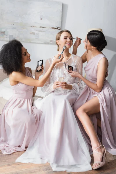 Multicultural women doing makeup to elegant bride in bedroom — Stock Photo