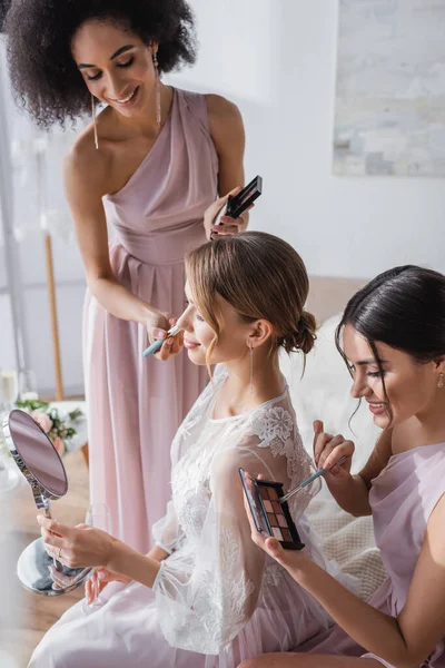 Jeune mariée regardant dans le miroir tandis que les femmes interracial appliquant le maquillage — Photo de stock