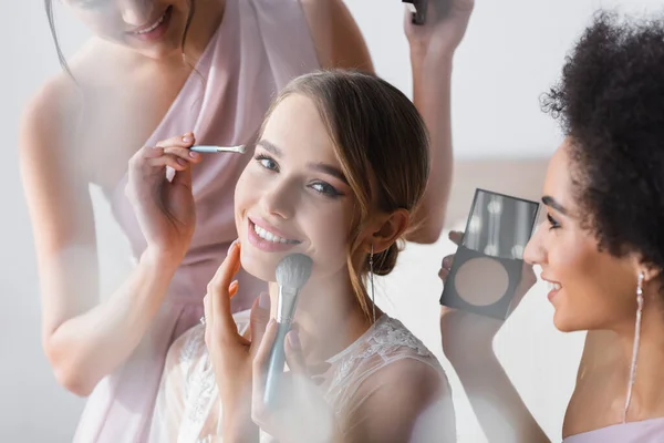 Bonita novia sonriendo a cámara mientras interracial las mujeres aplicando maquillaje, borrosa primer plano - foto de stock