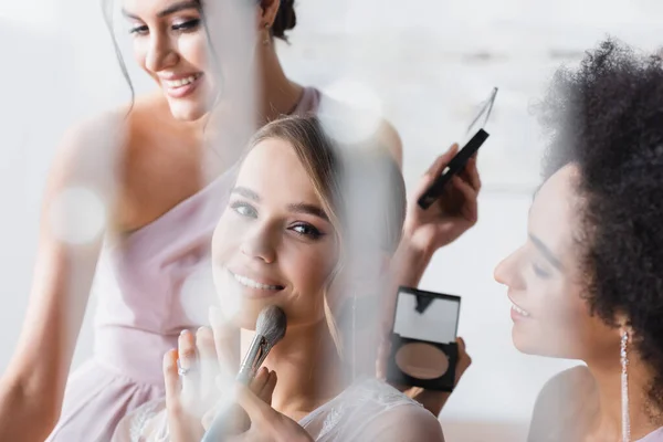Noiva feliz olhando para a câmera enquanto as mulheres inter-raciais aplicando maquiagem, foreground borrado — Fotografia de Stock